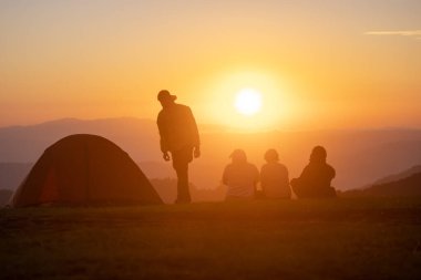 Bir grup arkadaş gece kampı sırasında çadırın yanında oturur ve güzel manzaralı gün batımını izlerler.