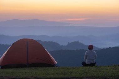 Gezgin, gece kampı sırasında çadırın yanında meditasyon yaparken açık havada macera seyahati için dağın tepesindeki güzel manzaralı günbatımına bakıyor.