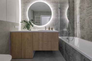 Modern minimalist bathroom interior design with wooden furniture, grey stone tiles, round mirror.  Aesthetic simple interior design concept.