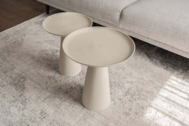 Modern oturma odasının içinde açık gri halı ve farklı boyutlarda iki yuvarlak desenli metal sehpa var.