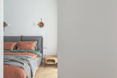A view through an open door into a bedroom interior with rust color linen and cushions on a bed, grey blanket, rattan bedside table and metal wall light.