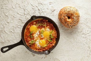 Shakshuka in cast iron pan with freshly baked bagel on white textured background. Fried eggs with tomato sauce. Shakshuka a traditional meal of the Jewish cuisine