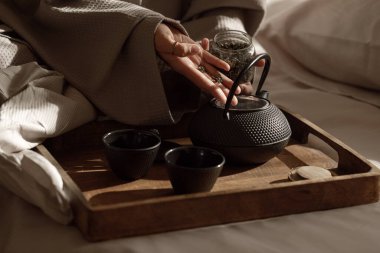 Yatağın üzerindeki ahşap tepside bitki çayıyla birlikte siyah dökme demir çaydanlık. Geleneksel Asya Çay Seti - Demir çaydanlık ve yeşil çay yapraklı seramik çay fincanları.