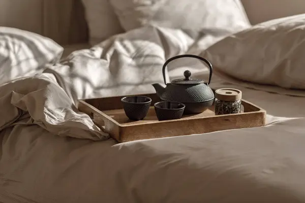 Black cast iron tea pot with herbal tea set up on wood tray on the bed. Traditional Asian Tea Set - iron teapot and ceramic teacups with green tea and tea leaves.