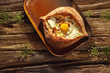 Geleneksel bir Khachapuri otantik bir tabağa dayanır, güneşli bir ışıkta, ahşap bir arka plana yaslanır, altın kabuğunu ve yapışkan peynirini ön plana çıkarır..