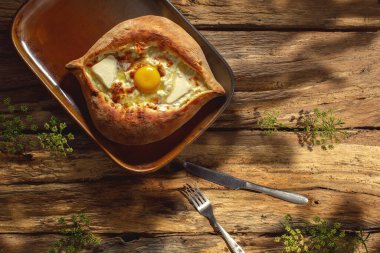 Geleneksel bir Khachapuri otantik bir tabağa dayanır, güneşli bir ışıkta, ahşap bir arka plana yaslanır, altın kabuğunu ve yapışkan peynirini ön plana çıkarır..