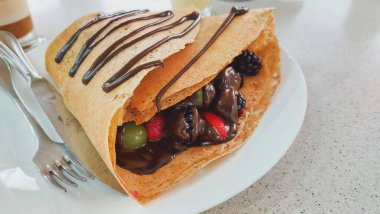 Çilek, böğürtlen, üzüm ve eritilmiş çikolata içeren, arka planda kapuçino kahveli, bir restoran masasında çikolatalı kırmızı meyve krepinin yakın çekimi..