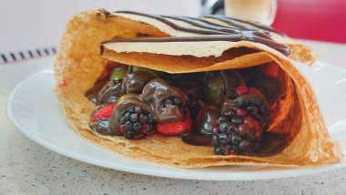 Çilek, böğürtlen, üzüm ve eritilmiş çikolata içeren, arka planda kapuçino kahveli, bir restoran masasında çikolatalı kırmızı meyve krepinin yakın çekimi..