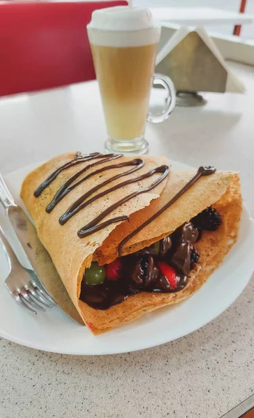 Vertical view of a red fruit crepe with chocolate, containing strawberries, blackberries, grapes and melted chocolate, with a cappuccino coffee in the background, at a restaurant table.