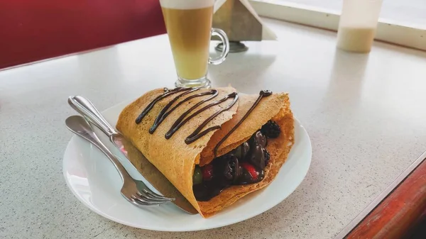 Horizontal view of a red fruit crepe with chocolate, containing strawberries, blackberries, grapes and melted chocolate, with a cappuccino coffee in the background, at a restaurant table.