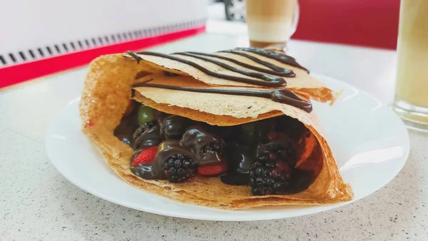 Close-up of a red fruit crepe with chocolate, containing strawberries, blackberries, grapes and melted chocolate, with a cappuccino coffee in the background, at a restaurant table.