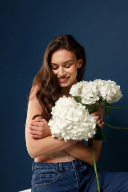 A happy fragile girl in blue jeans and a beige corset sits on a chair and holds a bouquet of hydrangeas in her hands. Blue background.