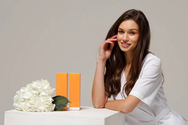 A beautiful young cosmetologist doctor in a white medical suit smiles at the camera and holds cosmetology products in her hands. Self care, beauty and cosmetology concept.