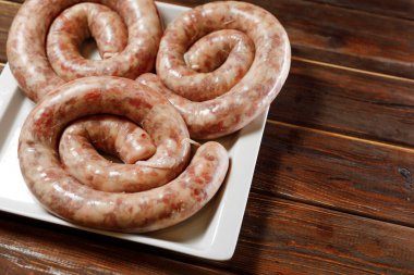 Resimde uzun sosisler görebiliyoruz. Ziyaretçi için kızartılmaya hazır. Kahverengi masadaki beyaz tabakta uzun sosisler var..