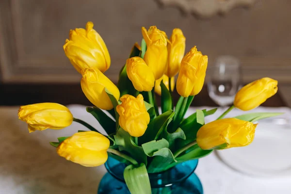 There is a vase on the table. We can see a vase with yellow flowers, three white plates and three glasses. We also can see a interesting mirror on the background. The vase is a beautiful.