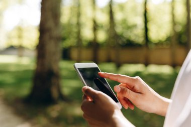 Akıllı telefonuna dokunan biri var. Arka planda bir park ve bir sürü ağaç görebiliyoruz. Adam yürürken telefonuyla bir şeyler yapıyor..