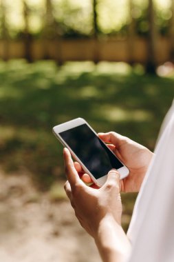 We see a person who is touching his phone. We also can see a park on the background and a lot of trees. The person is doing something in his phone while taking a walk. The weather is good. clipart