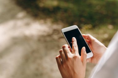 Telefonuna dokunan biri var. Arka planda bir park ve bir sürü ağaç görebiliyoruz. Yürüyüş yaparken telefonunda bir şey yapıyor. Hava güzel..