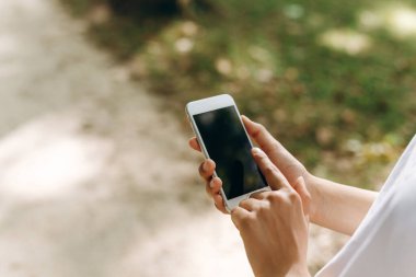 A human is holding the phone while walking. We also can see a park on the background and a lot of trees. The person is doing something in his phone while taking a walk. The weather is good. clipart