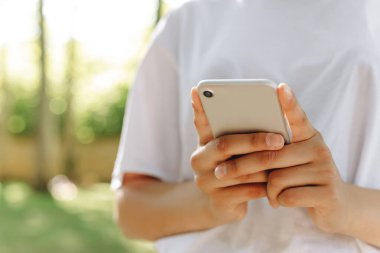 Güzel bir dişi yürüyor ve telefonla bir şeyler yapıyor. Arka planda bir park ve bir sürü ağaç görebiliyoruz. O, beyaz bir tişört giyiyor. Hava güzel..