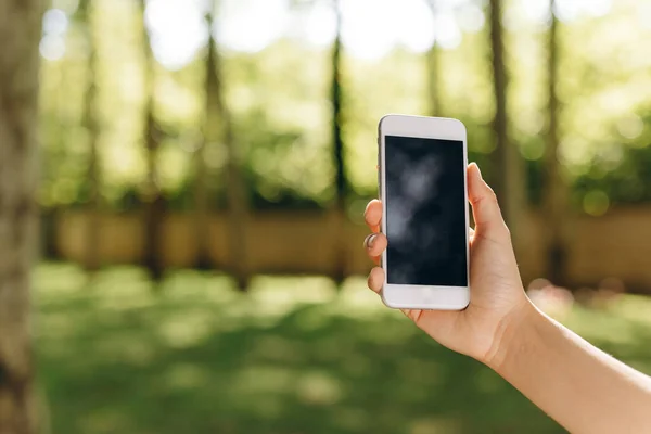 stock image A girl is holding smartphone while walking in the park. We also can see a park on the background and a lot of trees. The person is doing something in his phone while taking a walk. The weather is good.