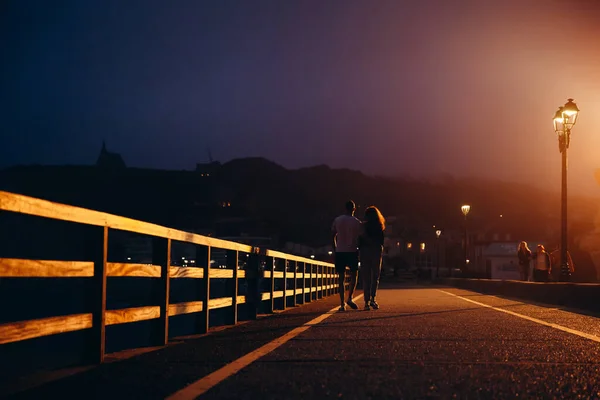 There is a brown railing made of wood on the bridge. It is evening or night right now. We can see a person walking towards the camera. There is a light on the right side of road. There is a couple.