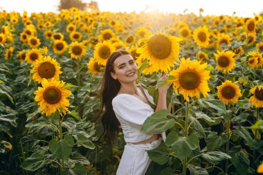Güzel kız rahatlıyor ve kameraya gülümsüyor. Çok koyu renk saçları var. Onun arkasında bir ayçiçeği tarlası var. Mutlu görünüyor. Arka planda bir gün doğumu görüyoruz..