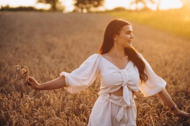 Gülümseyen yetişkin bir şeye bakarken sağ elinde buğday tutuyor. Beyaz bir gömlek giyiyor ve harika görünüyor. Güneş ışınlarını görebiliyoruz. Kızın arkasında büyük bir mısır tarlası var..