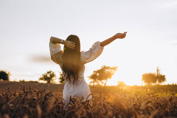 The amazing female is posing with her hands and looking at the sunset. She is wearing a white shirt and is looking amazing. We can see rays of sun. There is a big cornfield near the girl.