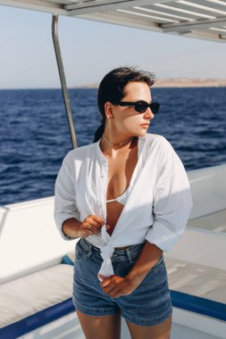 Deniz tatili ve teknede güneşin tadını çıkaran pozitif bir kadın. Neşeli, gülümseyen, beyaz bluzlu, şortlu ve güneş gözlüklü esmer kadın.,