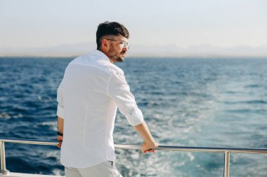 Bir yolcu gemisinin güvertesinde gülümseyen mutlu genç bir adam mavi gökyüzünün batımına doğru bakıyor..