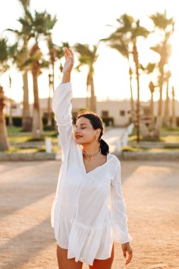Beyaz bir mayo içinde ışıl ışıl kar beyazı bir gülümsemesi ve denize karşı palmiye ağaçlarının yanında poz veren mutlu, ince bir kız. Yaz konsepti.