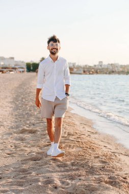Mavi gökyüzüne ve okyanusa tepeden bakan, harika bir tropik plajda poz veren sakallı yakışıklı bir adamın modaya uygun bir portresi klasik beyaz gömlek ve gözlüklerle günlük gri şort giymektedir..