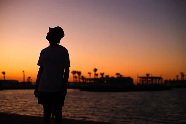 A silhouette of a young man with outstretched arms at the seafront with the background during sunset