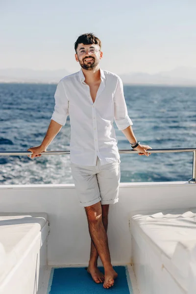 A stylish, young smiling happy man on the deck of a cruise ship on a sunny day and blue sky, looks into the camera. The concept of sea travel and recreation