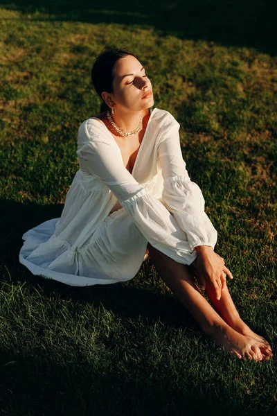 stock image A beautiful young happy girl enjoys a sunny day on the green grass, in a white dress. Vertical photo