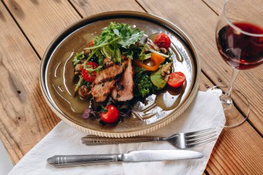 Bir tabak tabak, çatal bıçak ve bir bardak şarap var. Bir sürü yeşillik, birkaç domates ve kızarmış et görebiliyoruz. Her şey yağ ile doludur. İlginç ve lezzetli görünüyor..