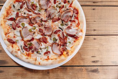 Pizzacının masasındaki büyük beyaz tabakta pizza yiyebiliriz. Lezzetli ve lezzetli görünüyor. Bir sürü et, sosis, salatalık ve peynir görebiliyoruz..