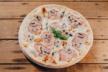 Ahşap masada mantar dilimli bir tabak pizza var. Dilimler lezzetli ve peynirli görünüyor. Ayrıca pizzada bir sürü sosis ve yeşil de var..