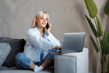 Güzel sarışın kız telefonda konuşuyor ve evde kanepede dizüstü bilgisayarla çalışıyor. Serbest meslek, iş, modern yaşam tarzı.