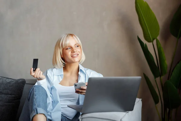 Evde otururken bilgisayarını ve telefonunu online alışveriş için kullanan kendine güvenen genç bir kadın, elinde kredi kartı tutan sarışın bir kız ve internet üzerinden ödeme yapan bir kız..