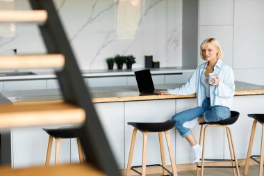 Genç sarışın bir kız evde, mutfakta dizüstü bilgisayarla çalışıyor. Doğal ışıklandırma var, beyaz tişört ve mavi gömlek giyiyor..