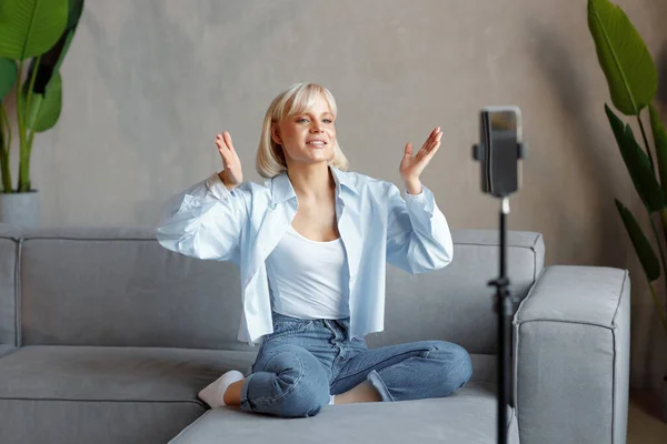 Stock image A young blonde girl is recording herself on the camera in the living room of her house, sitting on the couch. There is natural lighting, she is wearing a white T-shirt and a blue shirt.