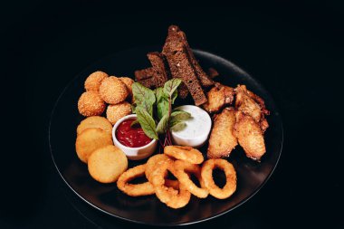 Bira seti. Kızarmış siyah ekmek, peynir topları, soğan halkaları ve kızarmış tavuk kanadı. İki çeşit soslu siyah bir tabakta servis ediliyor..