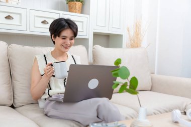 Rahat Asyalı serbest çalışan kadın öğrenci. Kanepede oturmuş bilgisayar ekranına bakıyor..