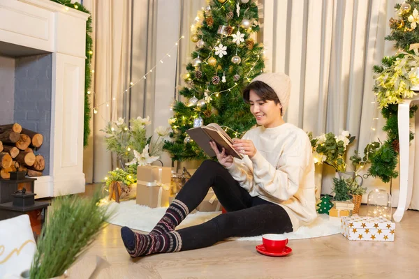 Asian woman in warm sweater and socks reading book sitting in room decorated for celebrating new year and christmas looking happy festive mood.