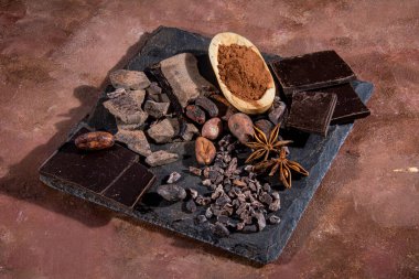 Kakao tozu, saf çikolata, kızarmış kakao çekirdekleri ve diğer malzemelerle birlikte bir levha üzerine doğranmış siyah çikolata tableti stüdyoda dokulu kahverengi arka plan taklit bir sıva üzerinde fotoğraflandı. Kaliteli kırsal görünümlü.