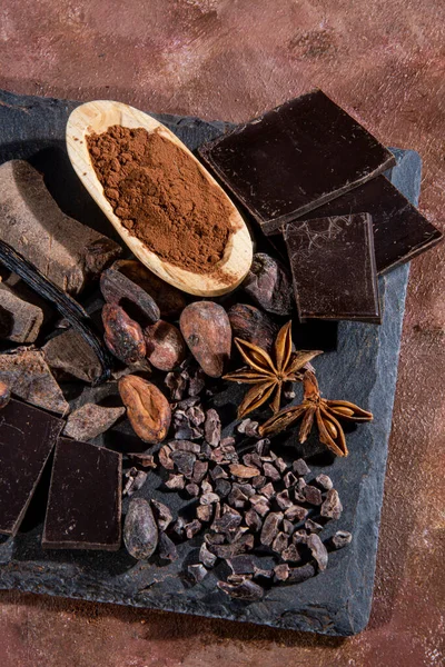 Black chocolate tablet chopped on a slate slab, accompanied by cocoa powder, pure chocolate, toasted cocoa beans and other ingredients, photographed in studio on a textured brown background imitating stucco. With vintage rustic look.