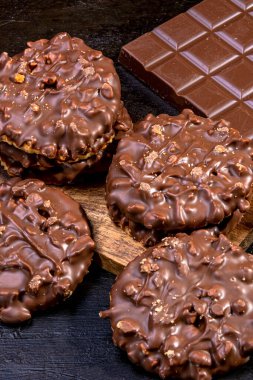Çikolata ve fındıklı kurabiyeler ahşap bir tepside, sütlü çikolata tabletinin yanında, fotoğrafçı tarafından elle boyanmış koyu bir arka planda izole edilmiş. Uzayı kopyala Çikolata ve fındıklı kurabiyeler ahşap tepside, sütlü çikolatanın yanında.