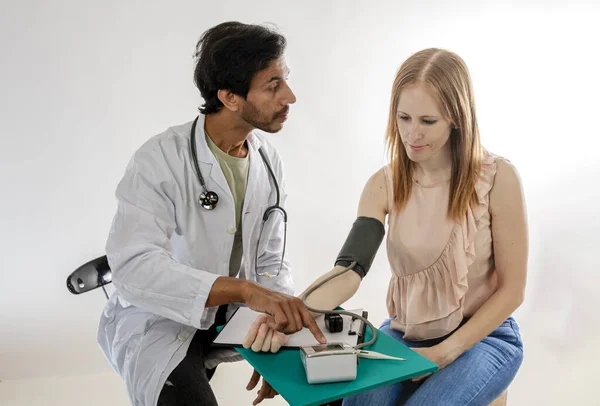 Asian doctor, from India, dressed in a white coat, measuring blood pressure on a redheaded European woman.